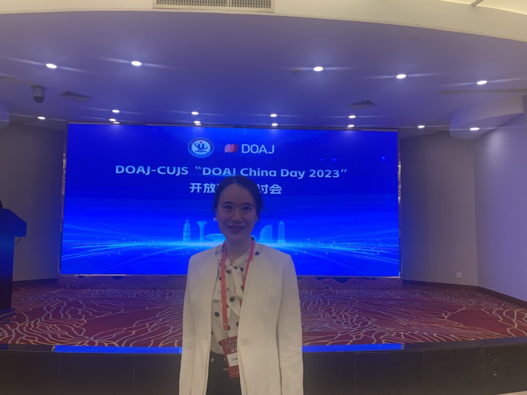Photo of Cenyu Shen standing in a conference room with DOAJ-CUJS "DOAJ China Day 2023" displayed on a large screen behind her